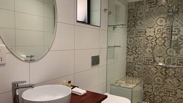 Een moderne badkamer met een grote, ovale spiegel, een witte wastafel, een douchecabine met glazen wanden en betegeling met een patroon.
