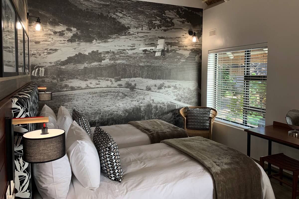 A bedroom with a large wall mural, a bed with patterned pillows, a chair, and a table with a glass.
