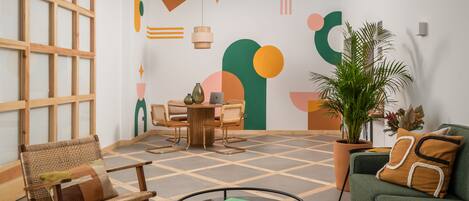 A modern living room with a wooden-framed window, a round glass coffee table, a green sofa, and a dining area with a wooden table and chairs.