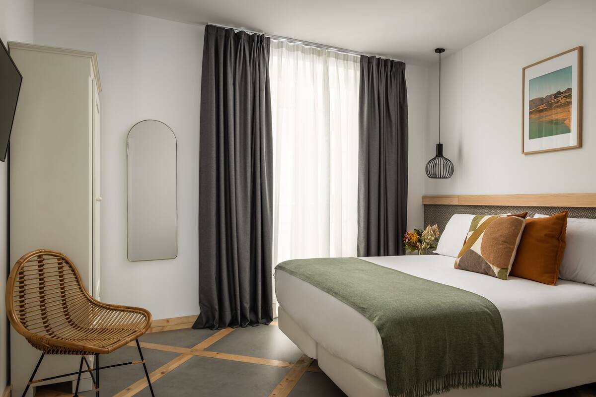 A hotel room with a bed, a chair, a mirror, and a painting on the wall.