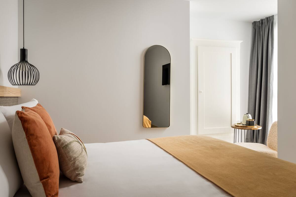 A modern hotel room with a bed, a bedside table, a mirror, and a window with curtains.