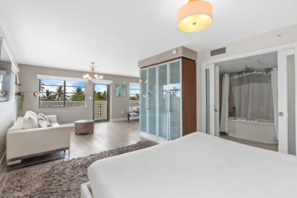 A modern bedroom with a large bed, a walk-in closet, a bathroom with a bathtub, and a view of palm trees through the windows. - Spacious Studio with Roof Top Pool & Bar (Miami Beach)