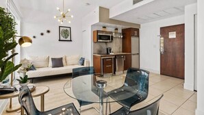 A modern living room with a glass dining table, a sofa, and a kitchen area with a microwave and oven.