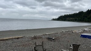 A pebbled beach with a wooden chair and a metal chair, a wooden log, and a small pile of debris. - Mckees Beach Cabin Steps From The Beach (Stanwood)