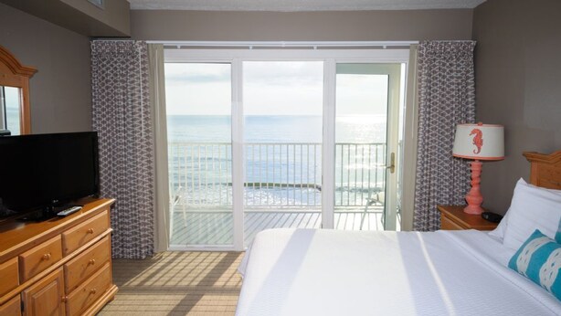 A hotel room with a bed, a TV on a wooden dresser, a nightstand with a lamp, and a balcony with a view of the ocean. - SRQ1 Enjoy unbeatable ocean views from the private balcony terrace, or oceanfront bedroom. (Kill Devil Hills)