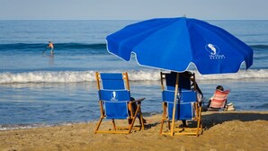 A beach scene with a blue umbrella, two beach chairs, and a person sitting under the umbrella. - SRQ1 Enjoy unbeatable ocean views from the private balcony terrace, or oceanfront bedroom. (Kill Devil Hills)