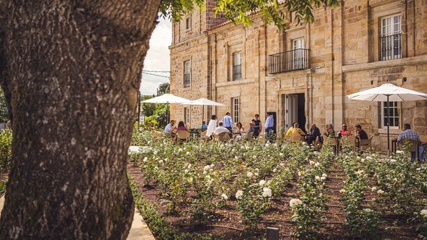 Un patio con edificios de piedra, mobiliario exterior y plantas de flores blancas.