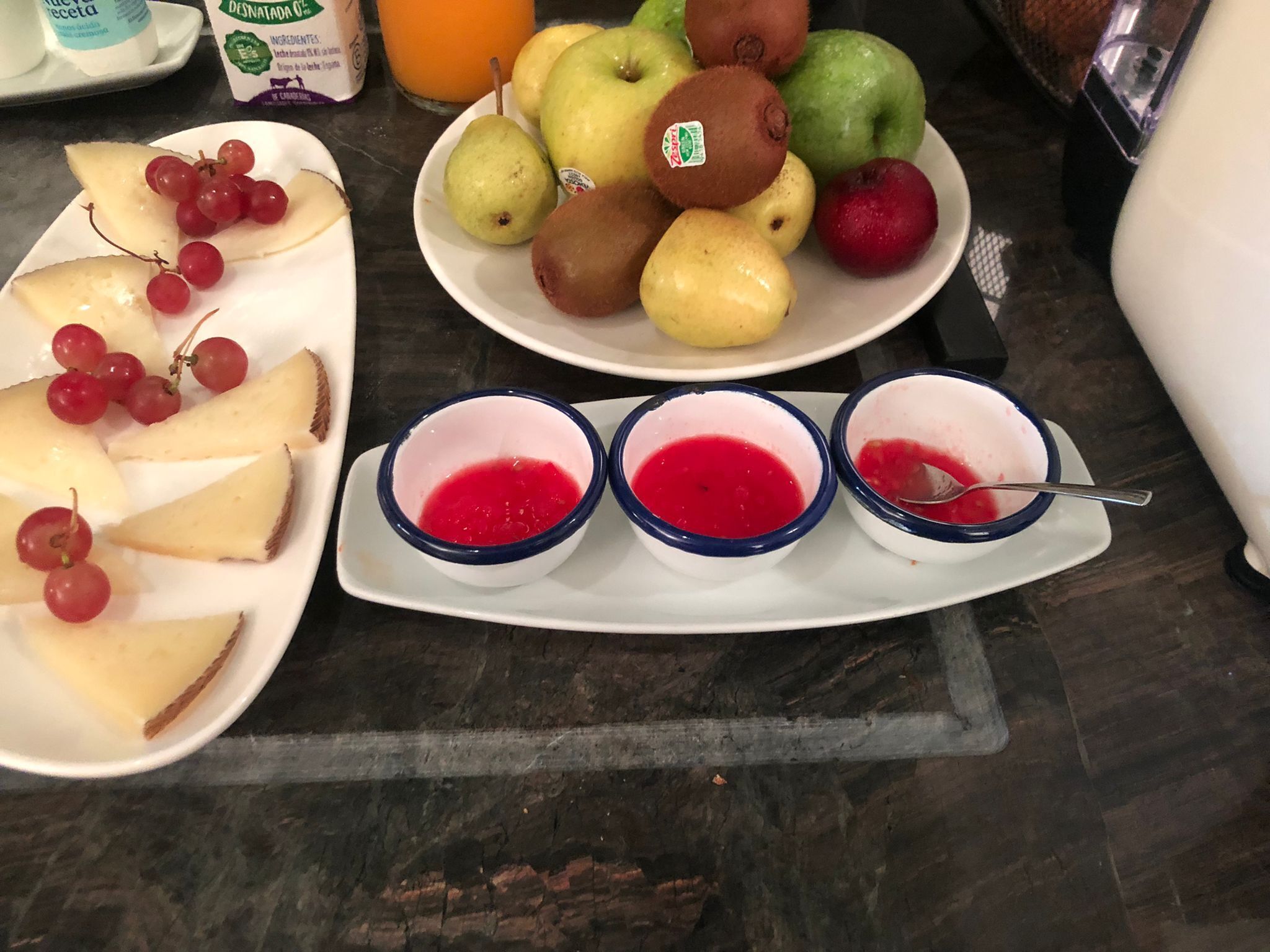 Una bandeja de frutas con uvas, peras y manzanas, y tres recipientes de mermelada roja sobre una encimera de mármol.