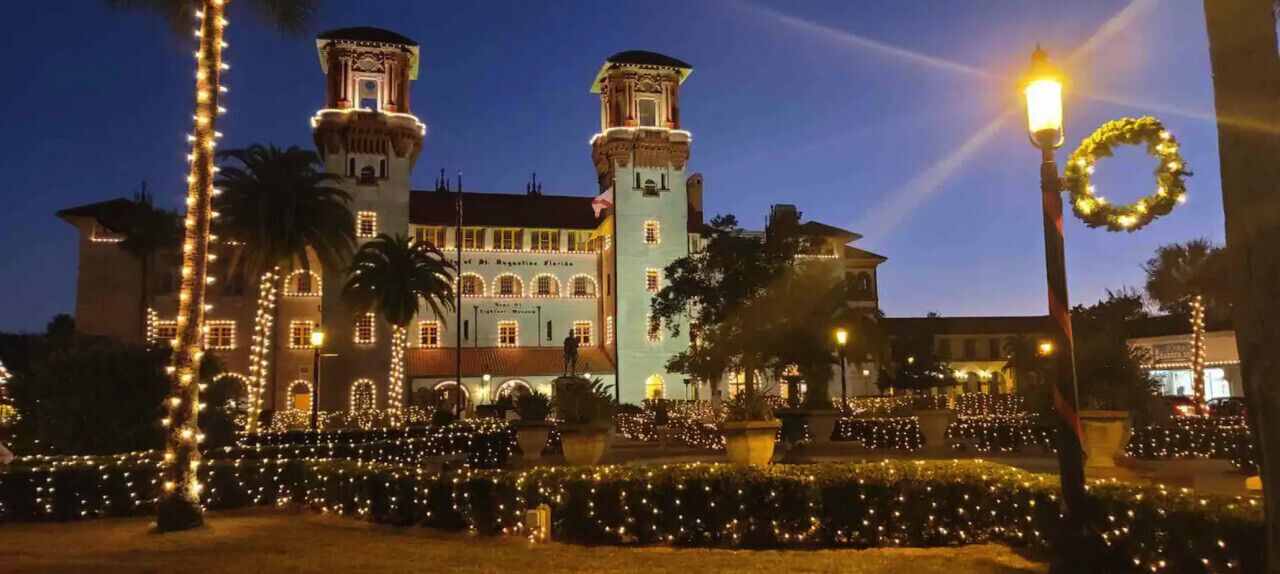 A historic building with two towers, surrounded by a well-lit garden with trees and lights.