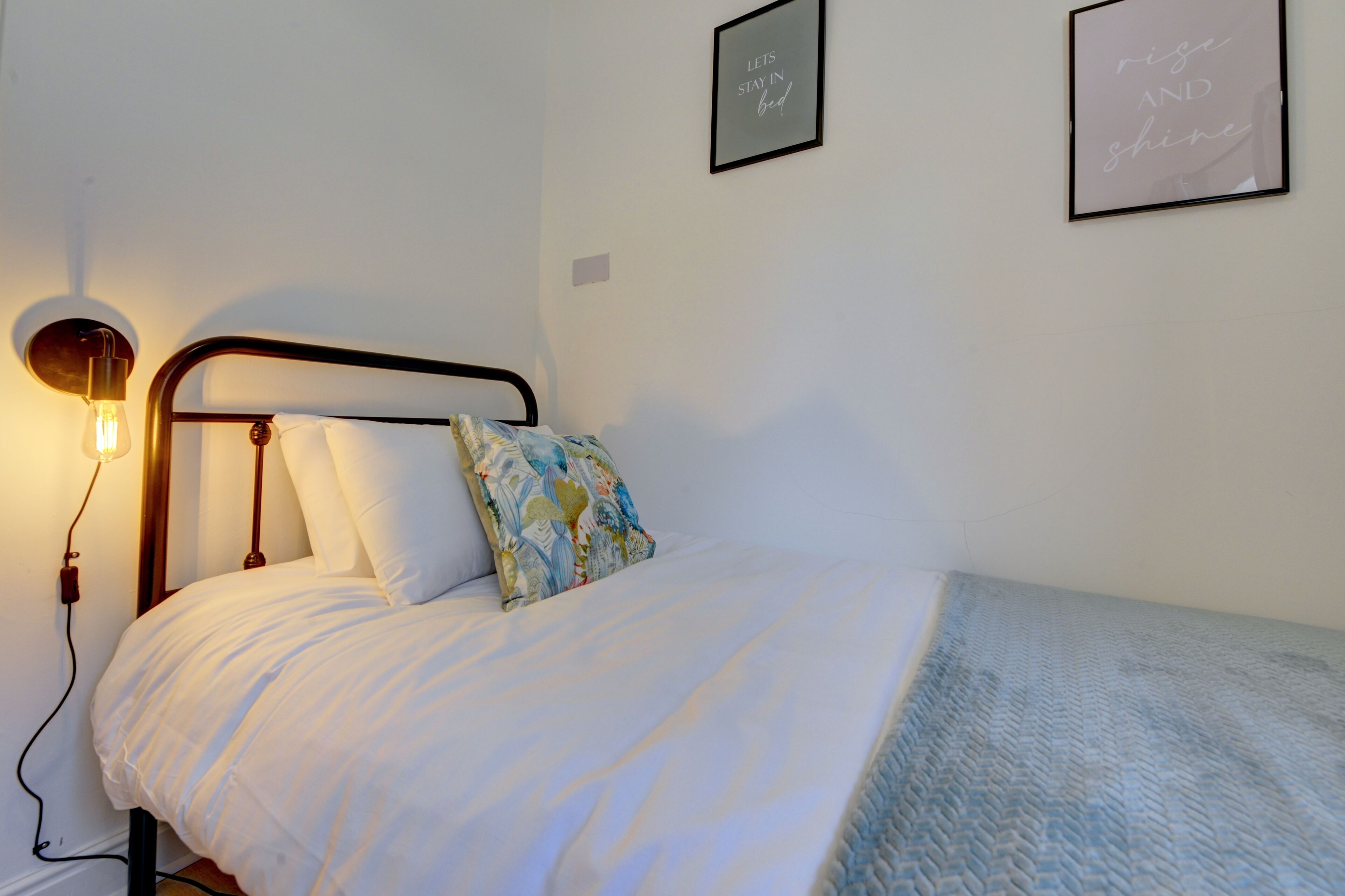 A neatly made bed with a white comforter and a colorful pillow, a bedside lamp, and two framed motivational quotes on the wall.