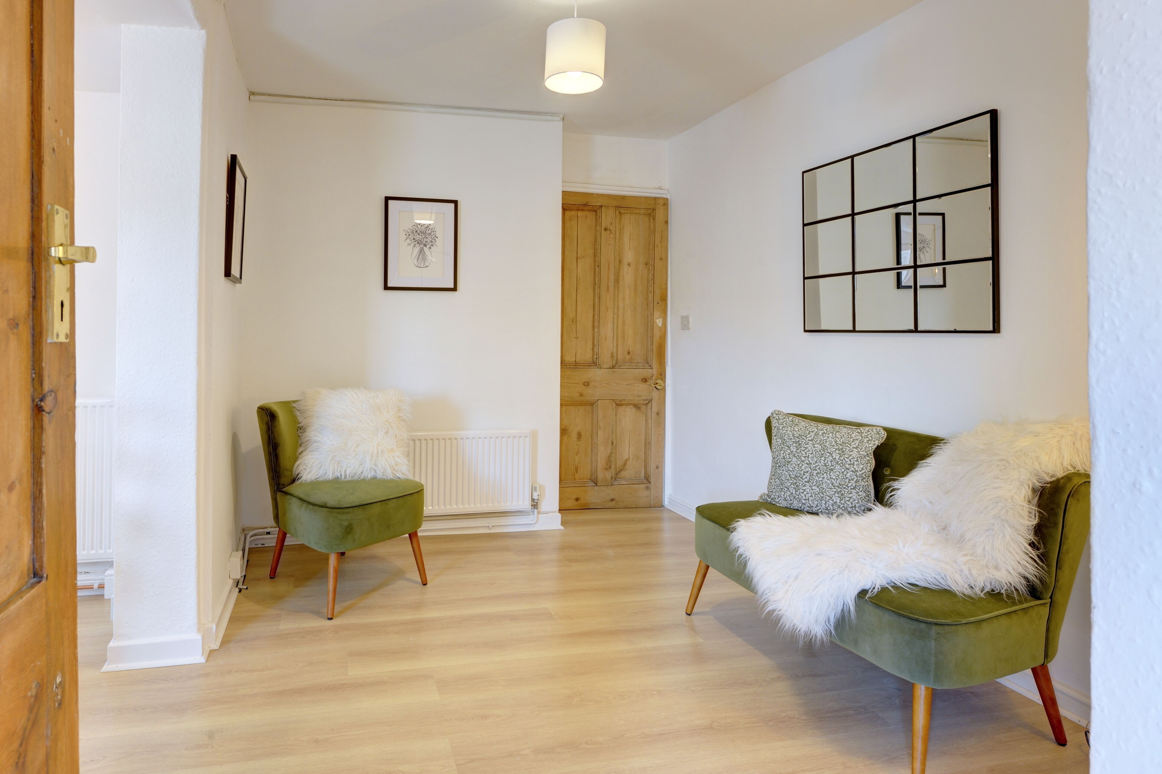A room with a wooden floor, two green armchairs with white fluffy throws, a wooden door, and a white radiator.