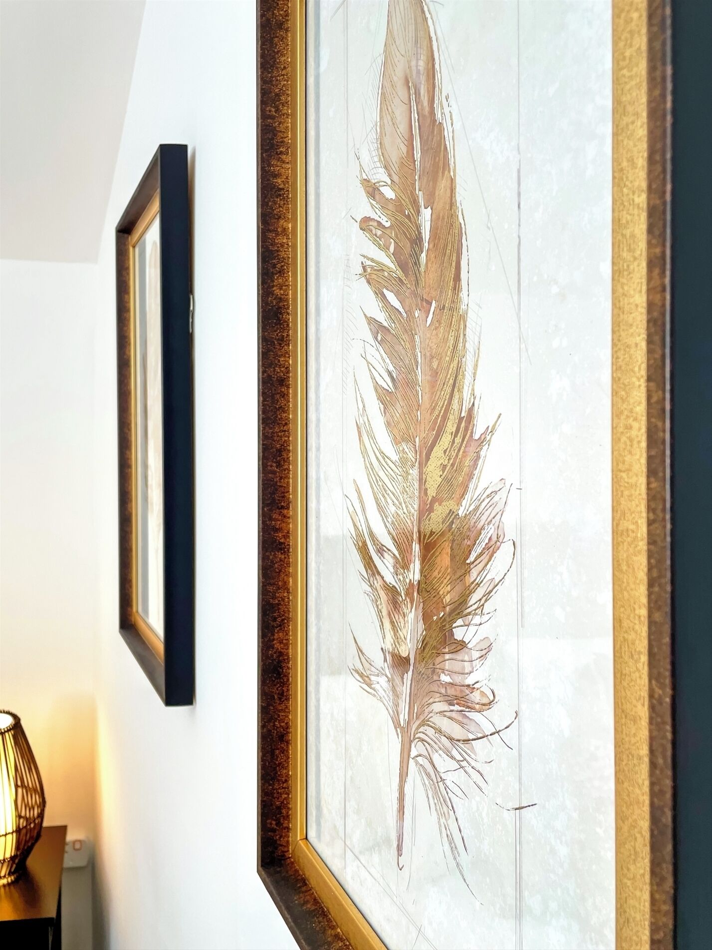 A framed feather artwork on a wall, a framed picture to the left, and a lamp on a table.