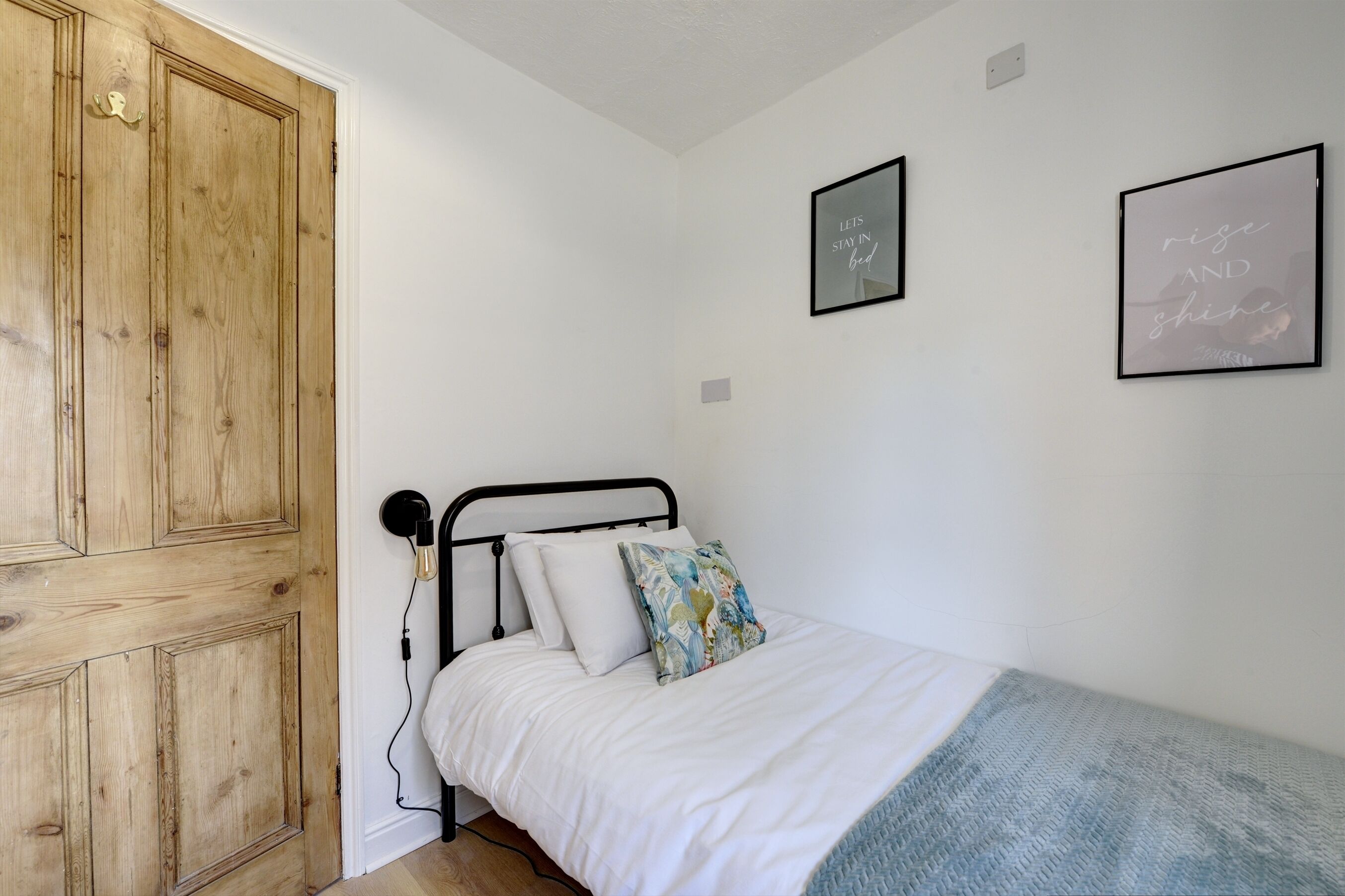 A bedroom with a wooden wardrobe, a bed with white bedding and a blue blanket, and two framed motivational quotes on the wall.