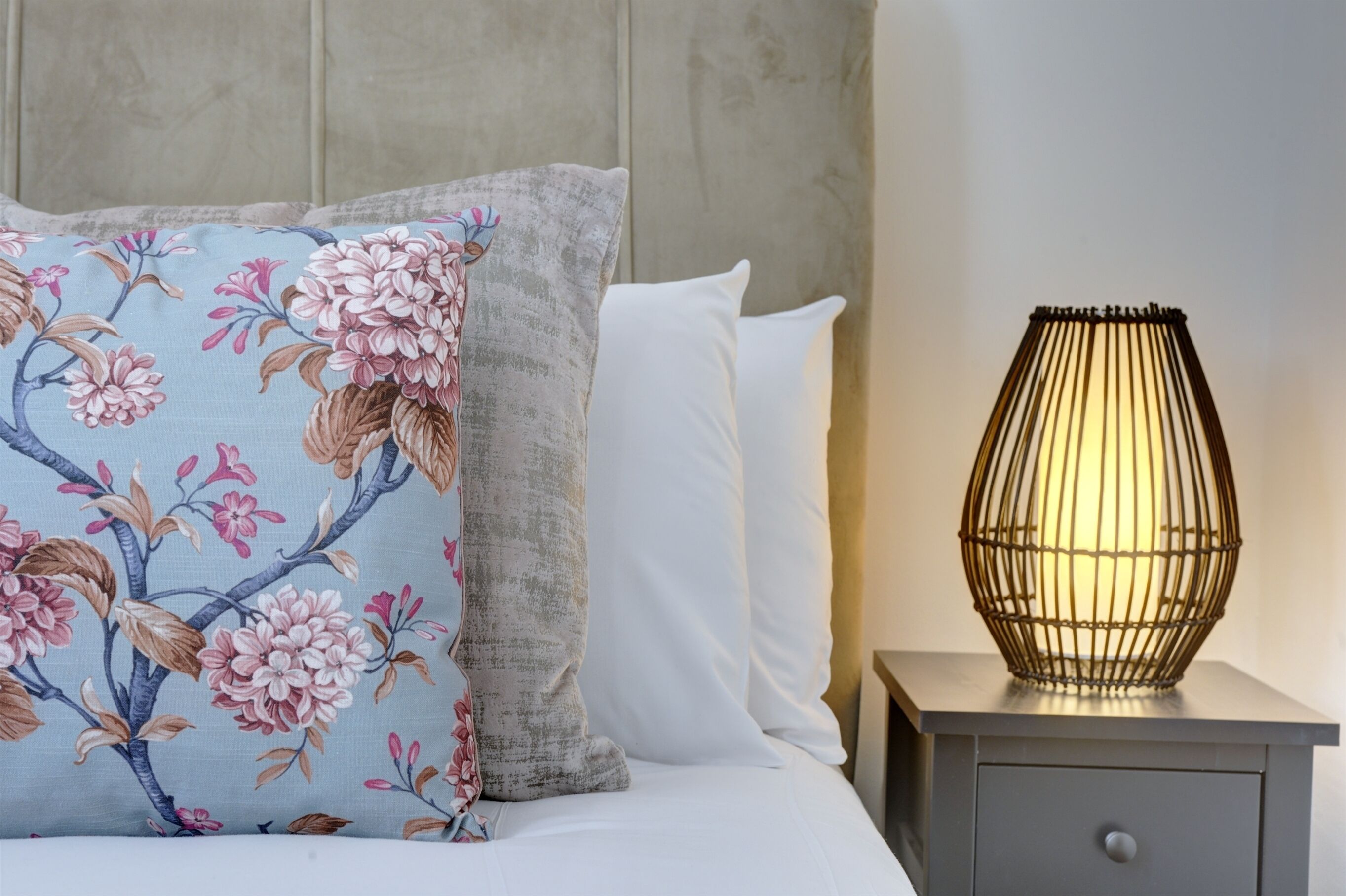 Ein Bett mit floralen Kissen, ein Nachttisch mit Lampe und ein grauer Beistelltisch.