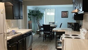 A kitchen with dark cabinets, a refrigerator, a stove, and a dining area with chairs and a table.