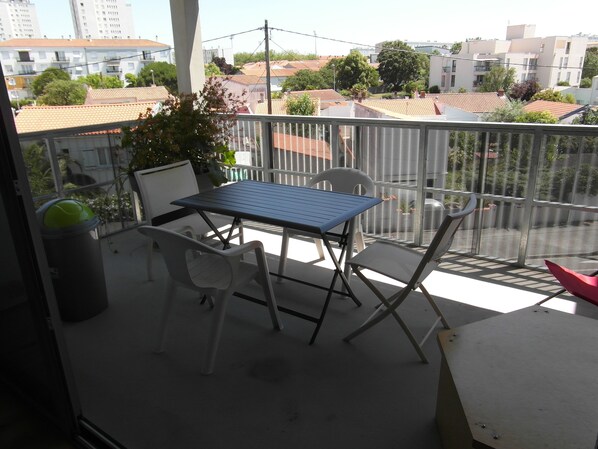 Un balcon avec une table et des chaises, une poubelle et une vue sur des immeubles et des arbres.