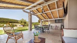 Un patio al aire libre cubierto con techo de madera, una mesa de comedor con sillas, una silla de mimbre con cojín, un banco de madera y un florero con flores.