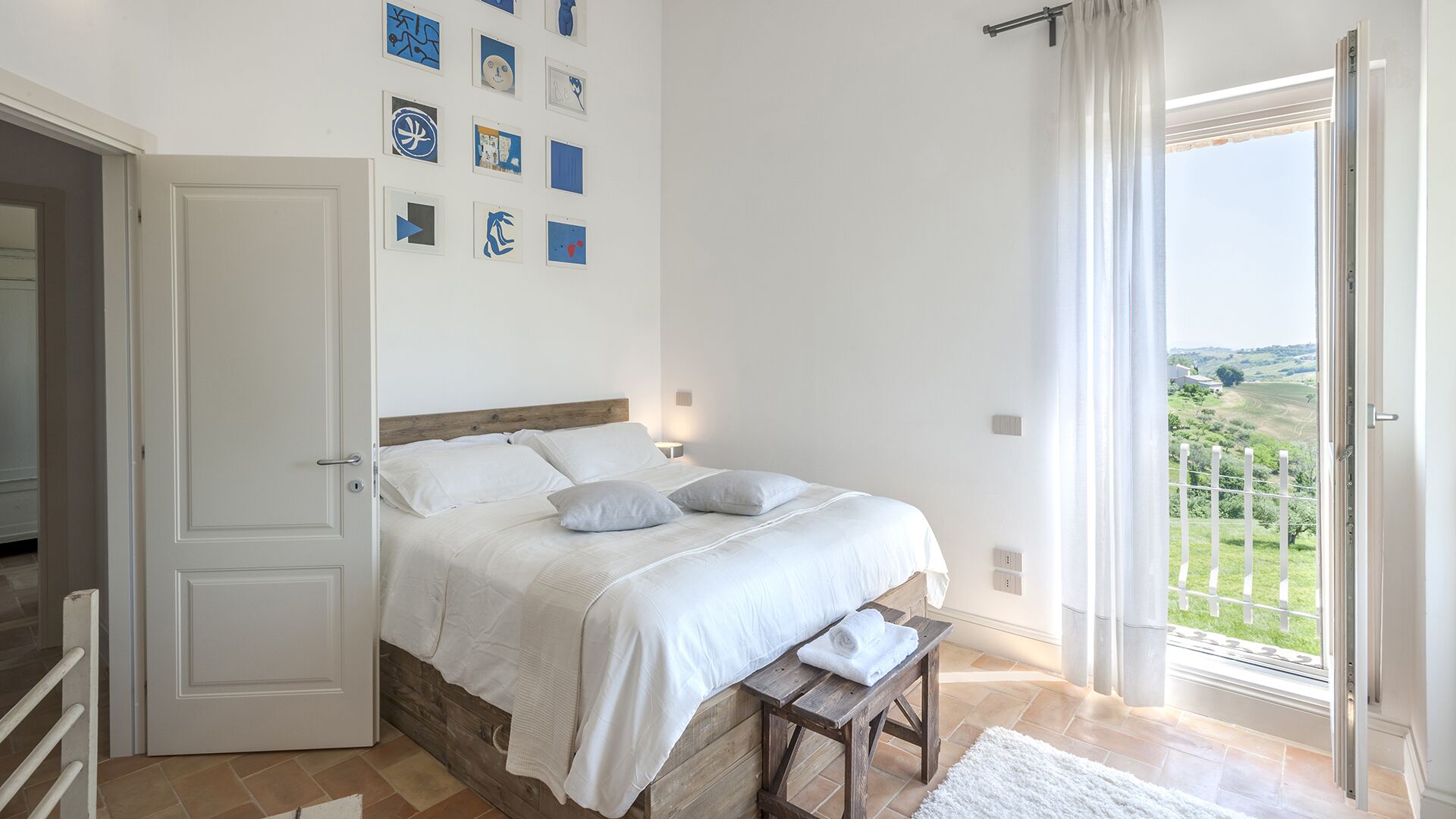 A bedroom with a bed, a bench, a view of the countryside, and a door.