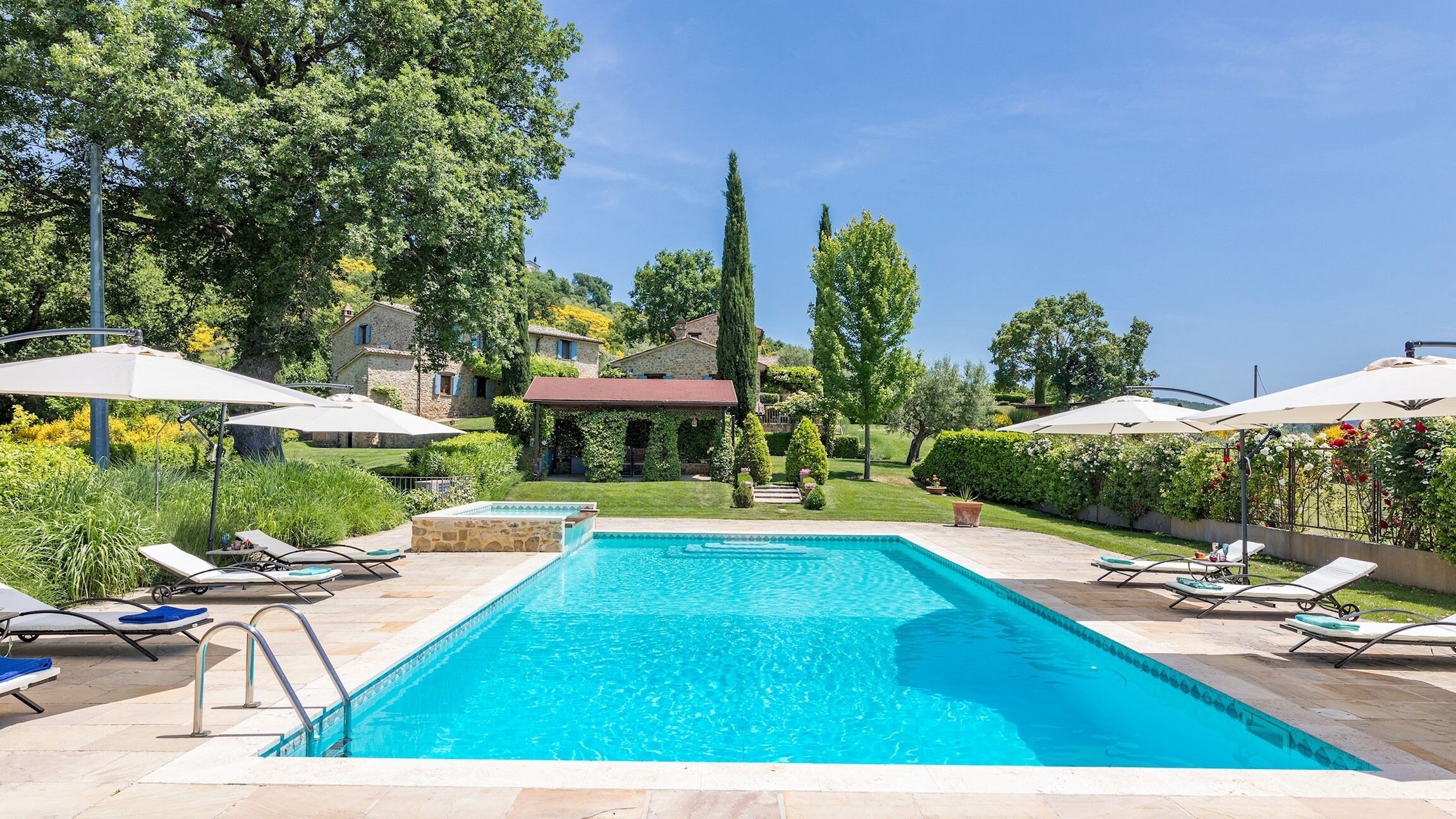 Une piscine rectangulaire bordée de pierres, entourée de chaises longues et de parasols, avec un bâtiment en pierre et des arbres en arrière-plan.