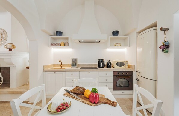 A modern kitchen with white cabinetry, a built-in oven, and a microwave. There is a dining table with a cutting board and vegetables.