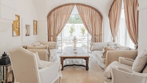 Ein geräumiges Wohnzimmer mit einem großen Tisch, zwei Sofas und Blick ins Freie durch große, mit Vorhängen drapierten Fenster.