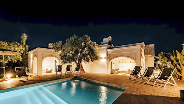 Ein Poolbereich bei Nacht mit klarem Pool, Liegestühlen und einem Gebäude mit Rundbogenfenstern.