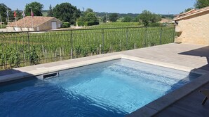 A swimming pool with a wooden deck, a stone wall, and a view of a vineyard.