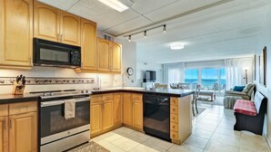 A modern kitchen with wooden cabinets, a microwave, stove, and dishwasher. There is a dining area with a view of the sky.