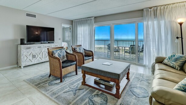 A spacious living room with a large flat-screen TV, wicker chairs, a beige sofa, and a view of the ocean through large sliding glass doors.