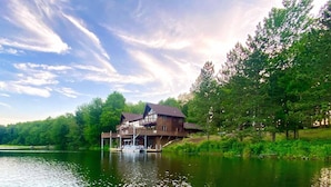 A large wooden house with a balcony, situated by a lake with boats docked nearby. - Custom built resort on private lake with kayaks, hiking, (Little Meadows)