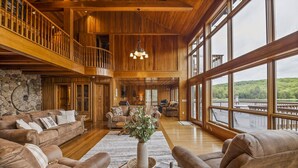 A spacious living room with a high ceiling, wooden beams, a stone wall, and large windows offering a view of a lake. - Custom built resort on private lake with kayaks, hiking, (Little Meadows)