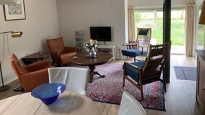 Una sala de estar moderna con una mesa redonda, sillones de cuero, una mesa de comedor con un tazón azul, un televisor y vistas a un jardín a través de amplios ventanales.