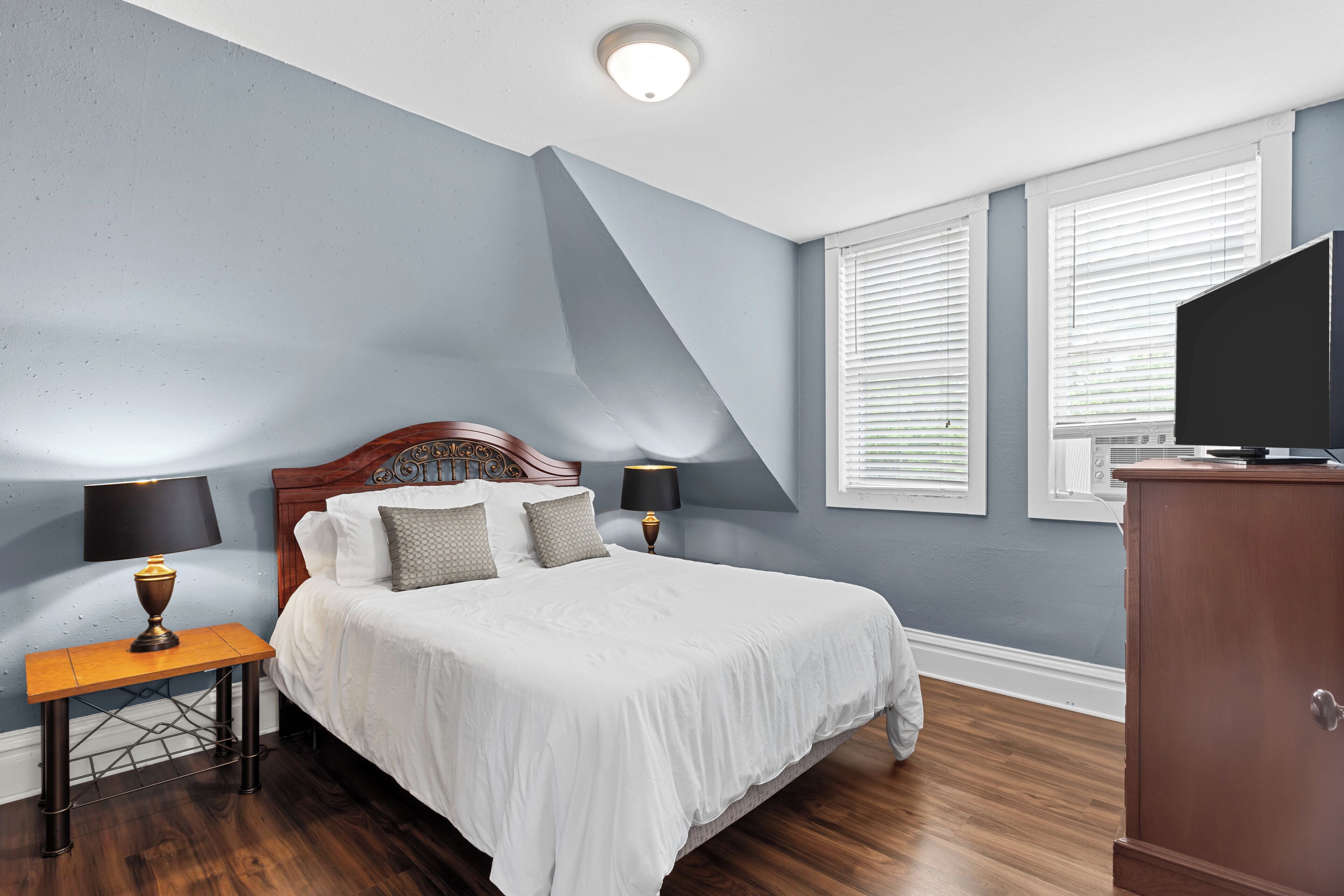 A bedroom with a large bed, two bedside tables with lamps, a wooden headboard, and a TV on a cabinet.