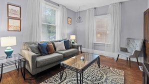 A living room with a sofa, coffee table, and armchairs, featuring large windows with white curtains and a patterned rug. - Historic 1908 Frenchtown Home (Saint Charles)