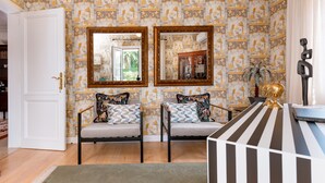 A room with patterned wallpaper, two chairs with cushions, a mirror, a desk with a laptop, and a door leading to another room.