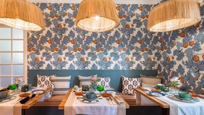 A dining area with a patterned wallpaper, wooden tables, and woven lampshades.