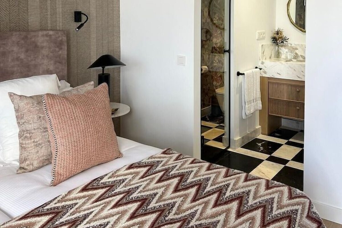 A modern bedroom with a bed, a nightstand, a lamp, an air conditioning unit, a bathroom with a sink and mirror, and a patterned rug.