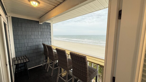 Un balcon avec des fauteuils en osier et une petite table, donnant sur une plage où l’on peut admirer les vagues.