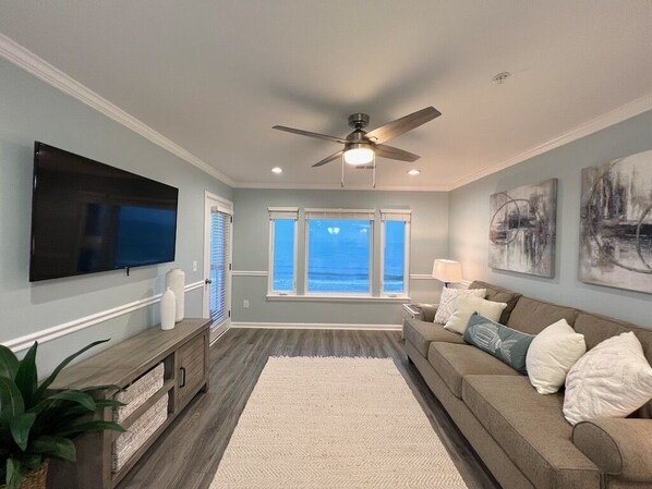 A living room with a flat-screen TV, a ceiling fan, a large window with a view, and a couch with pillows.