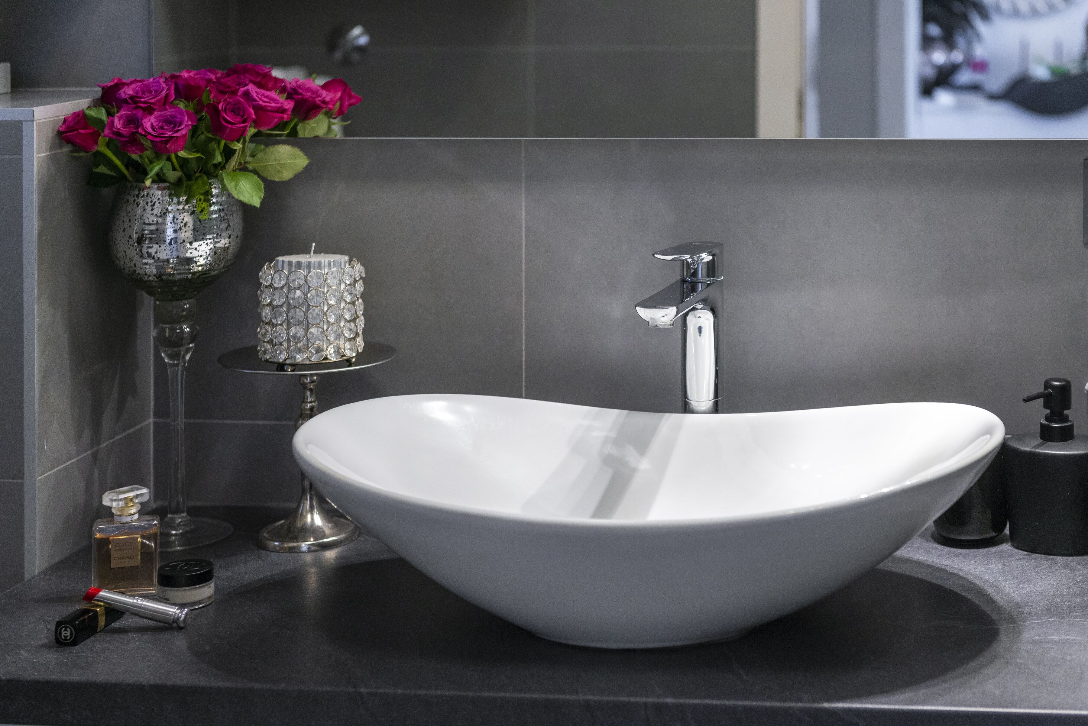 A modern bathroom with a large white vessel sink, a mirror, a vase with pink roses, and a small table with a candle and perfume bottles.