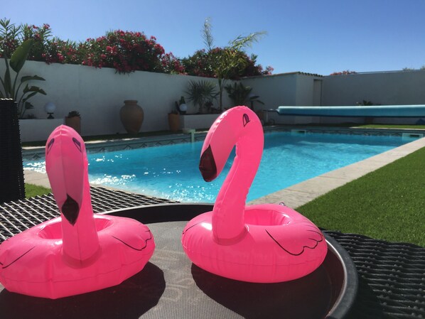Two pink flamingo inflatable floats by a pool with a clear blue sky and a white wall with red flowers in the background.