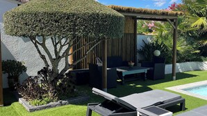 Un jardín trasero con césped artificial, una pérgola, sillones de descanso, una piscina y diversas plantas.