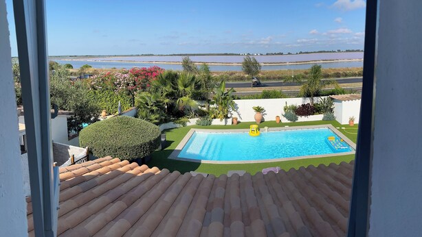 Vista desde un balcón sobre una piscina, con un cielo azul despejado y una masa de agua en la distancia.