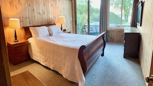 A bedroom with a wooden headboard, a bed with white bedding, two bedside lamps, a wooden nightstand, a bench, a dresser, and a large window with a view of greenery. - Heros Hideaway, private 1acre flat grassy yard, majestic sunsets (South Hero)