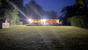 A house with a porch and outdoor seating area, surrounded by a grassy yard and trees. - Heros Hideaway, private 1acre flat grassy yard, majestic sunsets (South Hero)