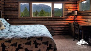 Ein Schlafzimmer in einer Blockhütte mit einem Bett, einem Schreibtisch und zwei Fenstern mit Blick auf die Berge.