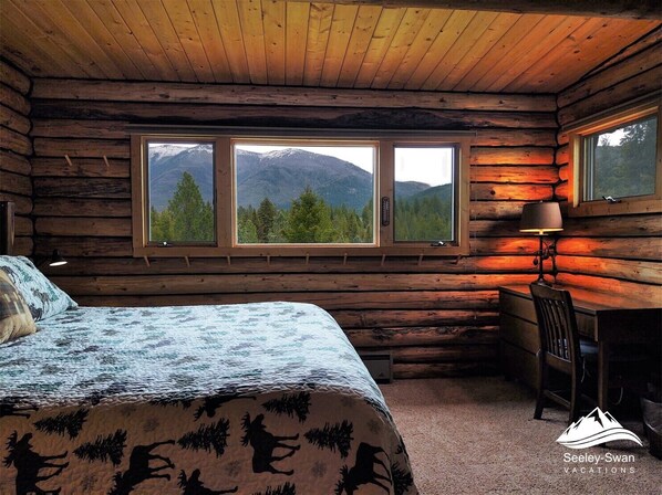 Une chambre aménagée dans un chalet en rondins, avec un lit, un bureau et deux fenêtres donnant sur une vue sur les montagnes.