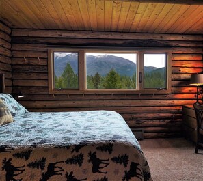 Une chambre aménagée dans un chalet en rondins, avec un lit, un bureau et deux fenêtres donnant sur une vue sur les montagnes.