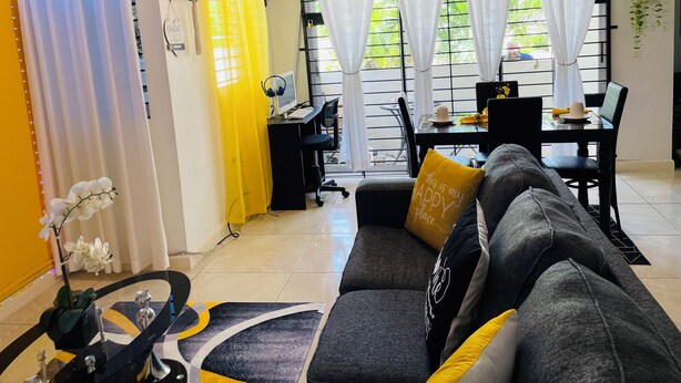 Ein Wohnzimmer mit einer Couch, einem Esstisch mit Stühlen und einem Schreibtisch mit einem Stuhl. Vor den Fenstern hängen Vorhänge, und eine Pflanze ist sichtbar.
