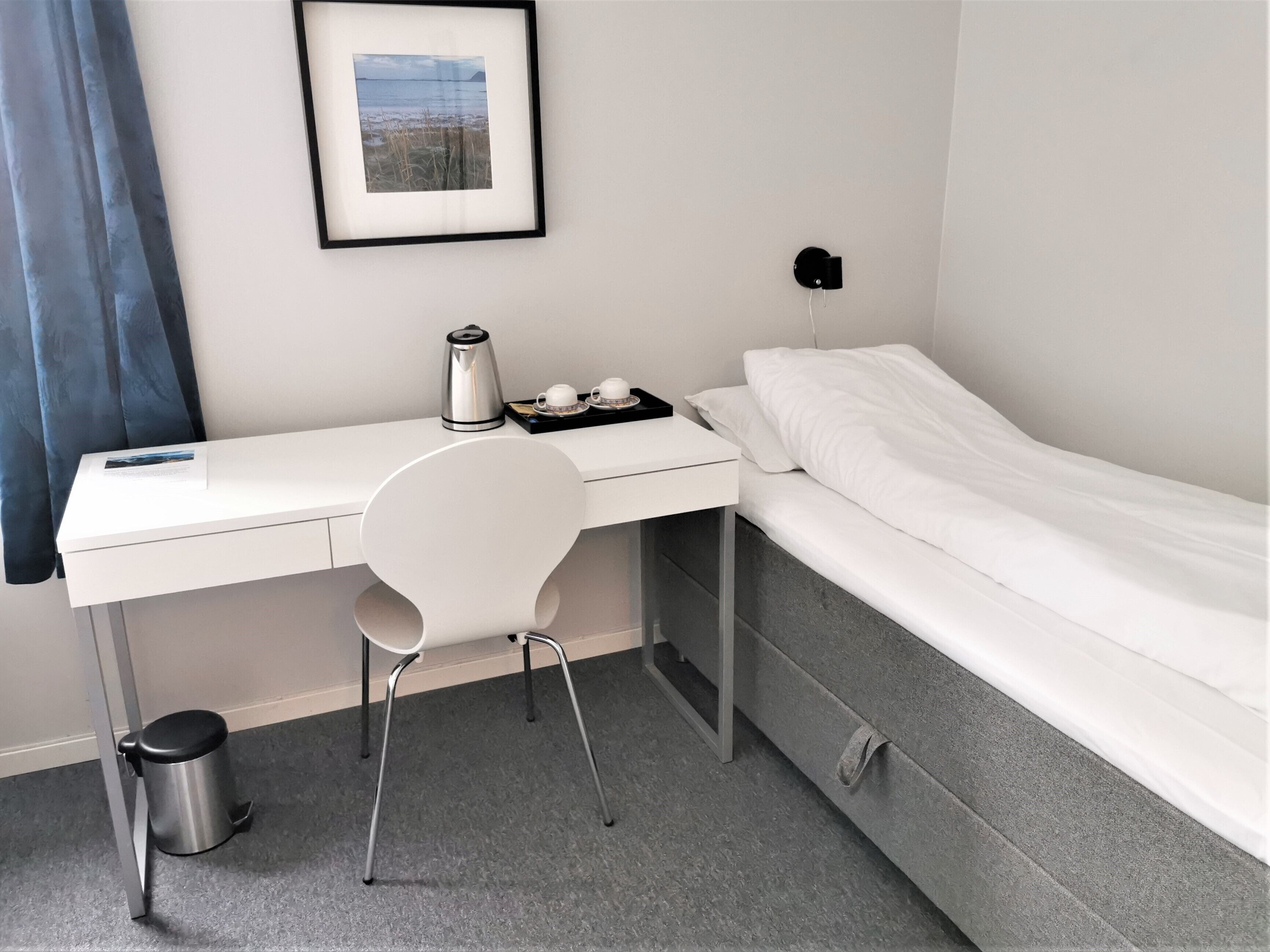 A compact hotel room with a white desk, a chair, a bed, and a framed picture on the wall.
